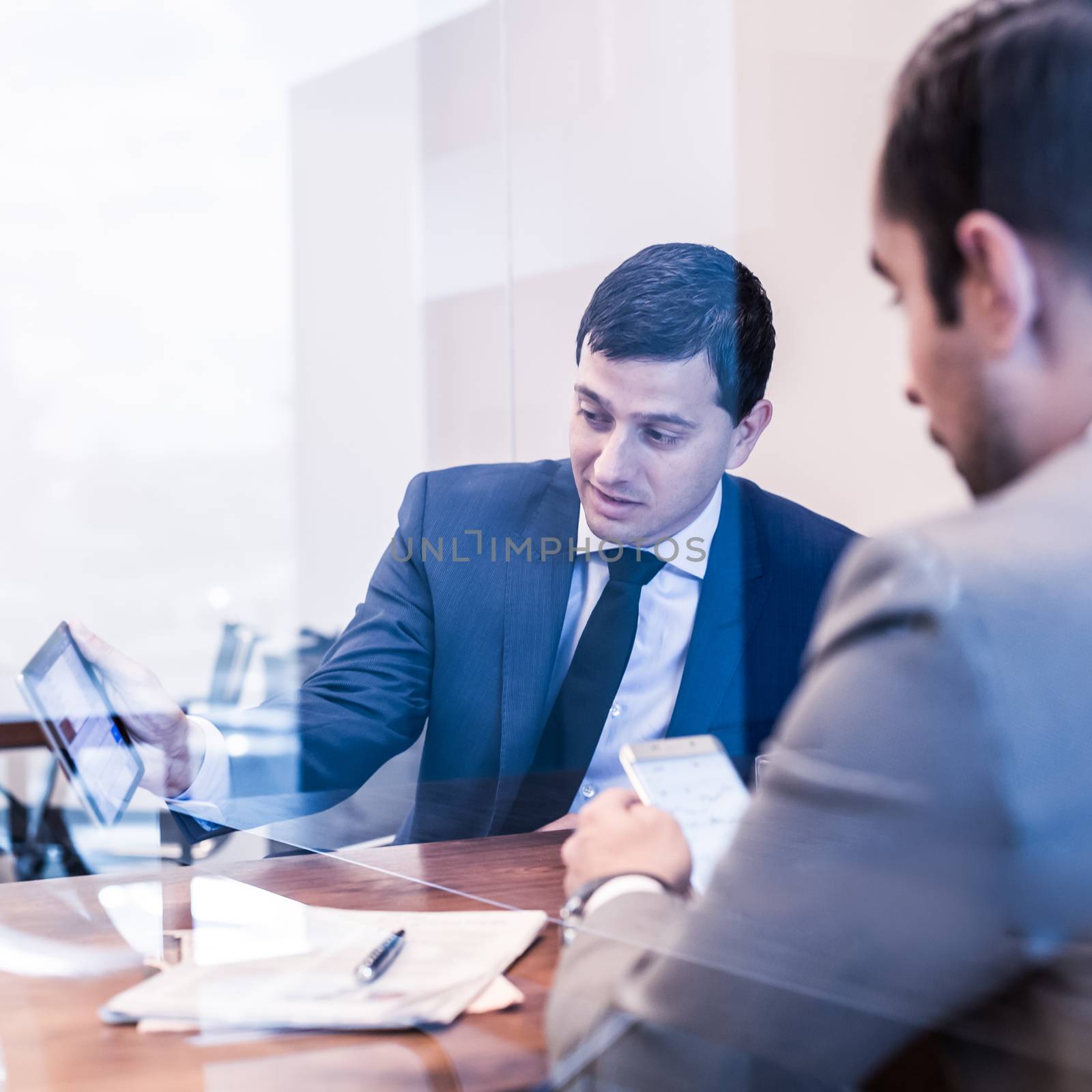 Through the window view of two young businessmen using tablet and smart phone devices at business meeting in corporative office.