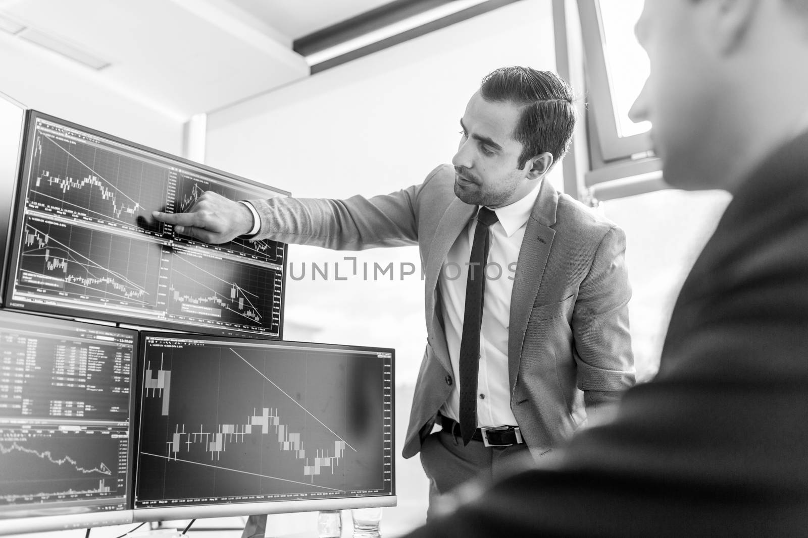 Businessmen trading stocks online. Stock brokers looking at graphs, indexes and numbers on multiple computer screens. Colleagues in discussion in traders office. Black and white image.