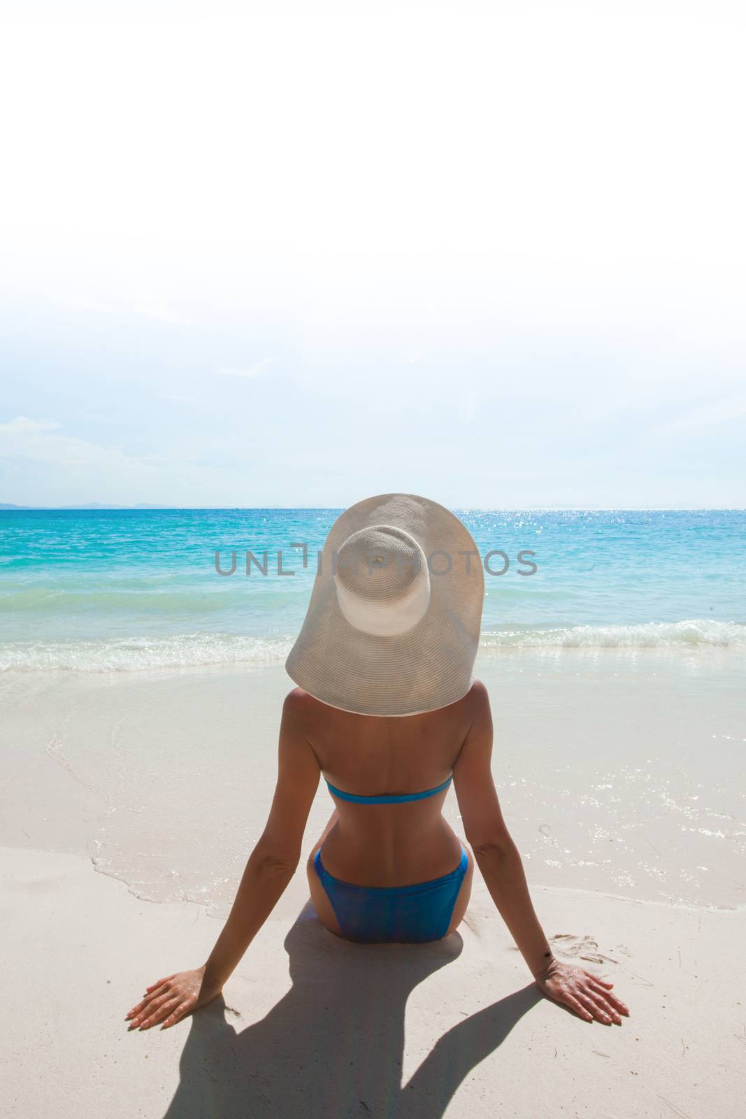 Woman in bikini and sunhat sitting on beach and looking at sea