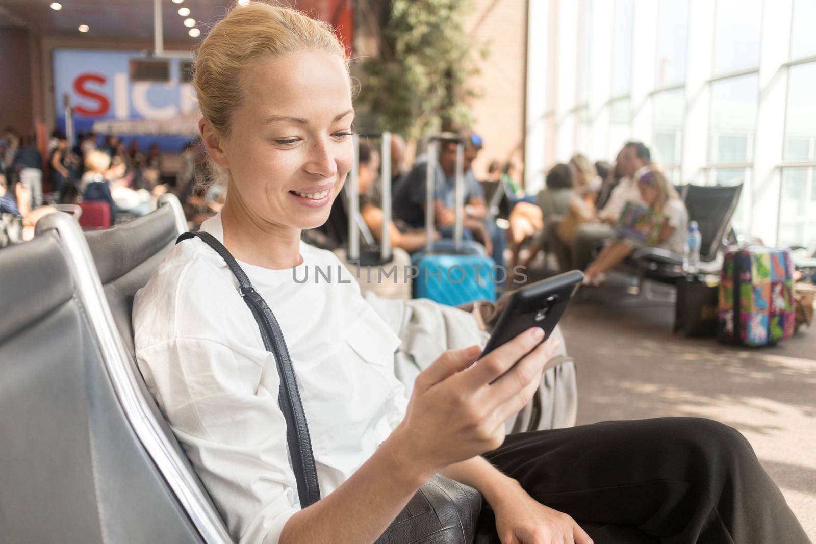 Casual blond young woman reading on her mobile phone while waiting to board a plane at the departure gates at the airport terminal. Travel concept.