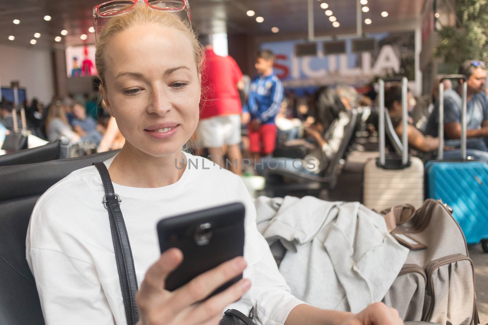 Casual blond young woman reading on her mobile phone while waiting to board a plane at the departure gates at the airport terminal. Travel concept.