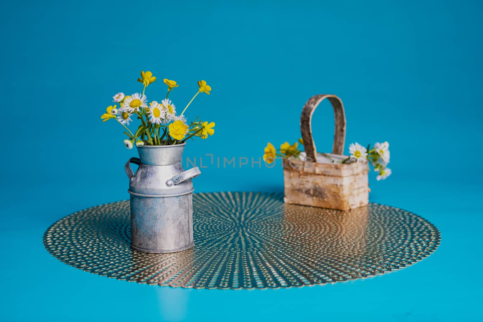 Hairy buttercup and prairie fleabane wildflowers in a miniature milk can and backet. Sitting on a placemat against a teal blue background.