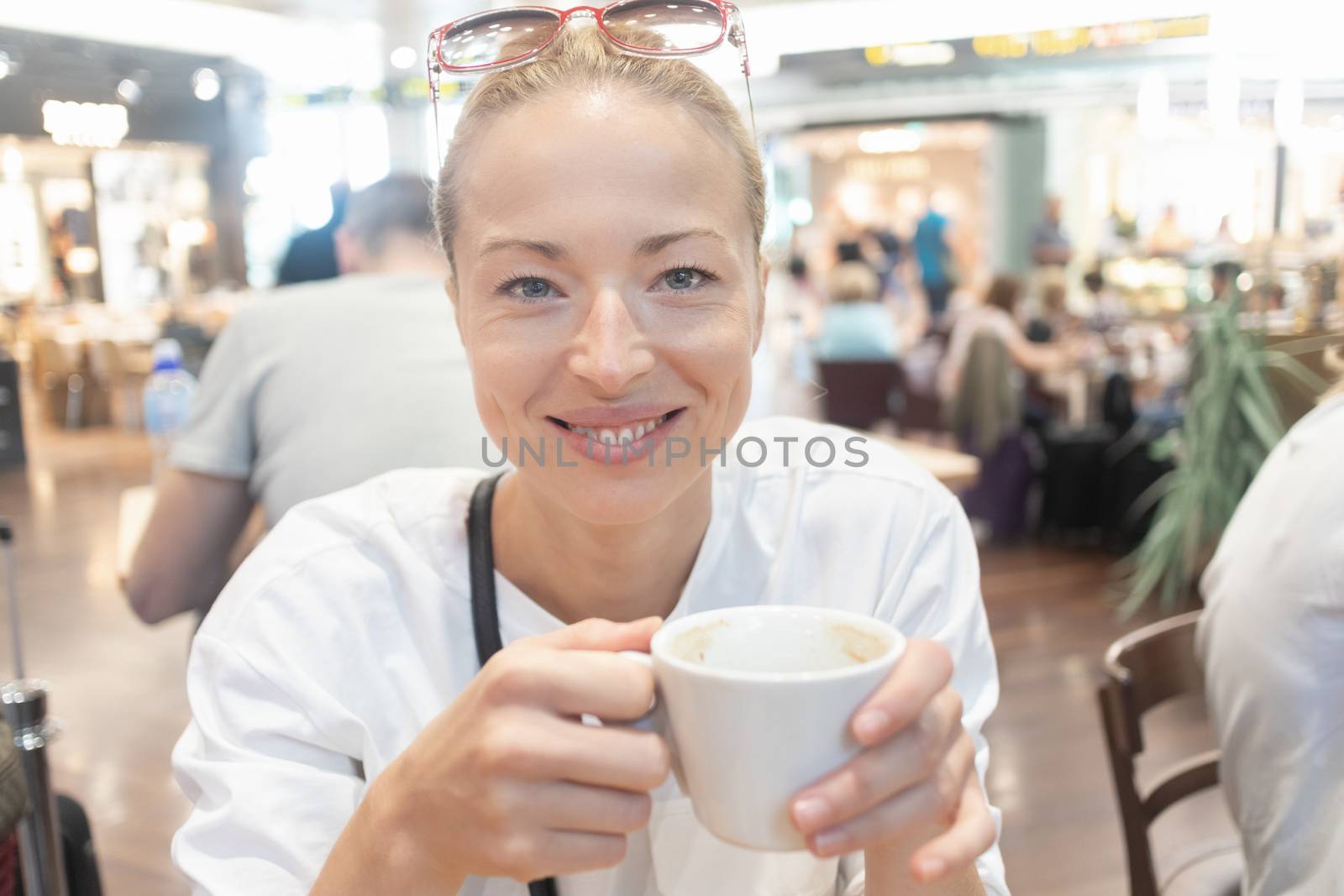 Portrait of a casual young blond woman having a cup of coffee, sitting and waiting in cafe indoors of an airport, station, food court or shopping mall.