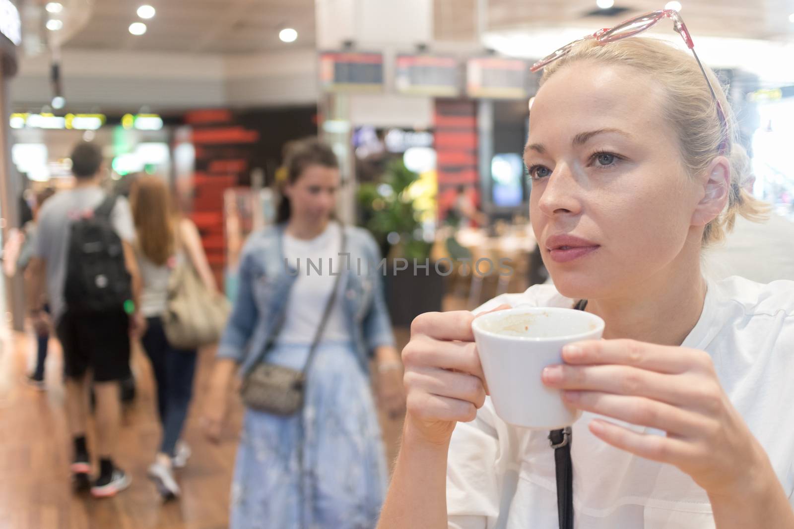 Portrait of a casual young blond woman having a cup of coffee, sitting and waiting in cafe indoors of an airport, station, food court or shopping mall.