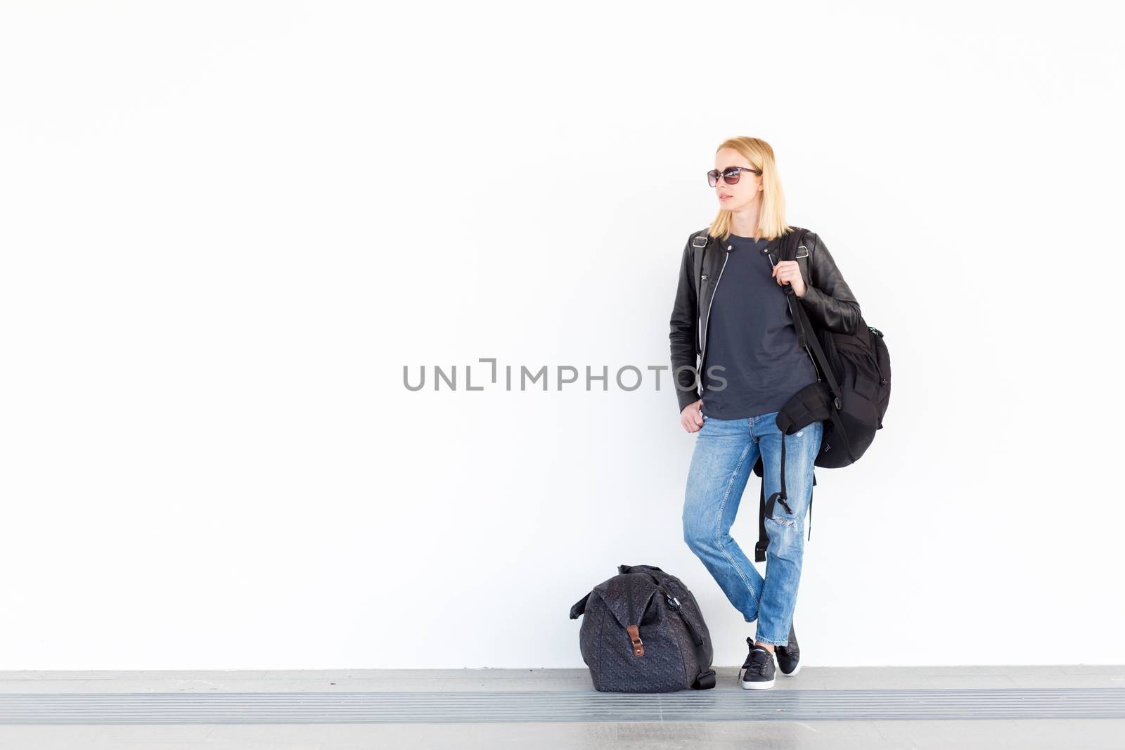 Fashionable young woman standing and waiting against plain white wall on the station whit travel bag by her side. by kasto