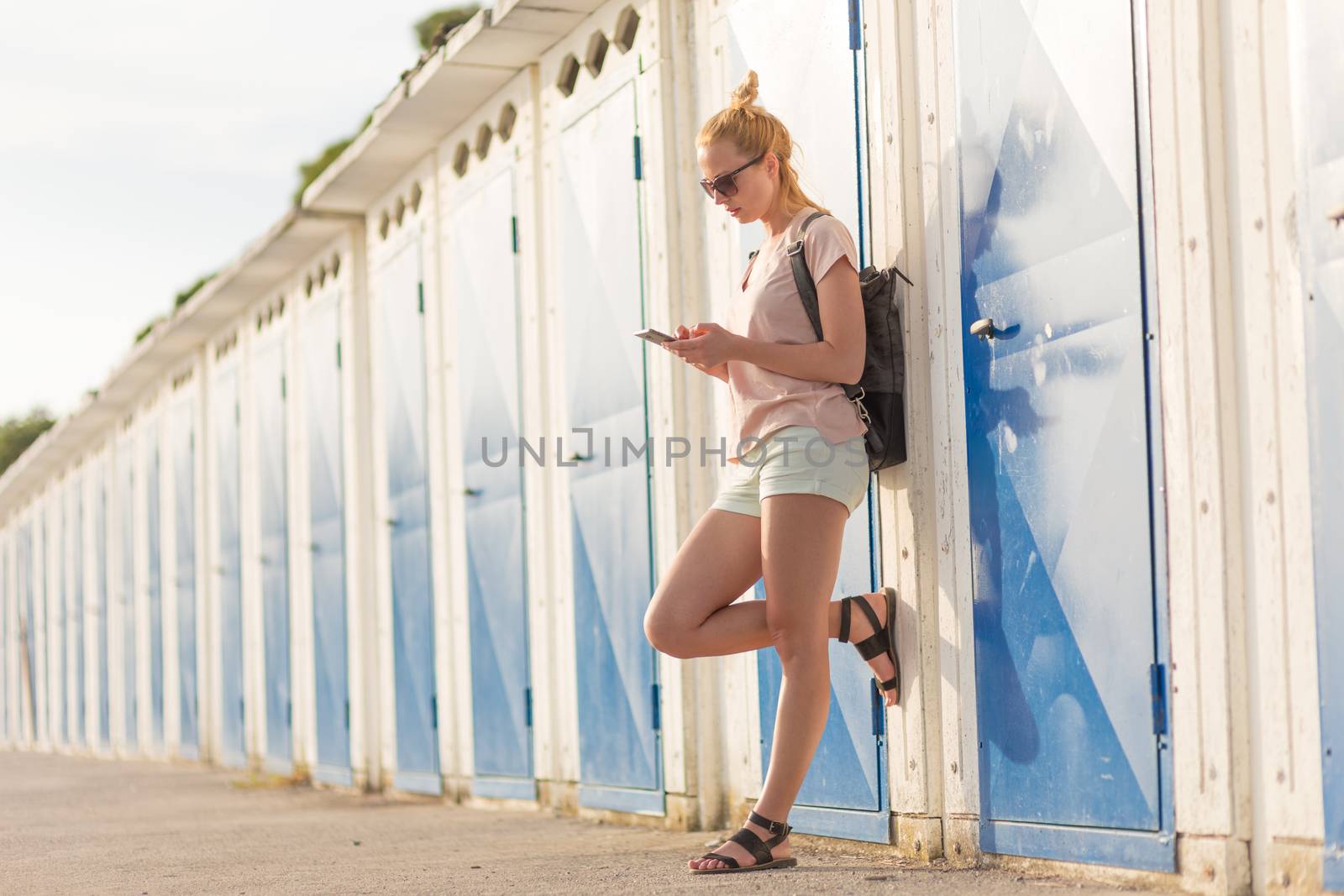 Blonde young female traveler wearing summer style clothing, using mobile phone, leaning against retro blue beach dressing rooms at summer time vacation in Sistiana, Italy.