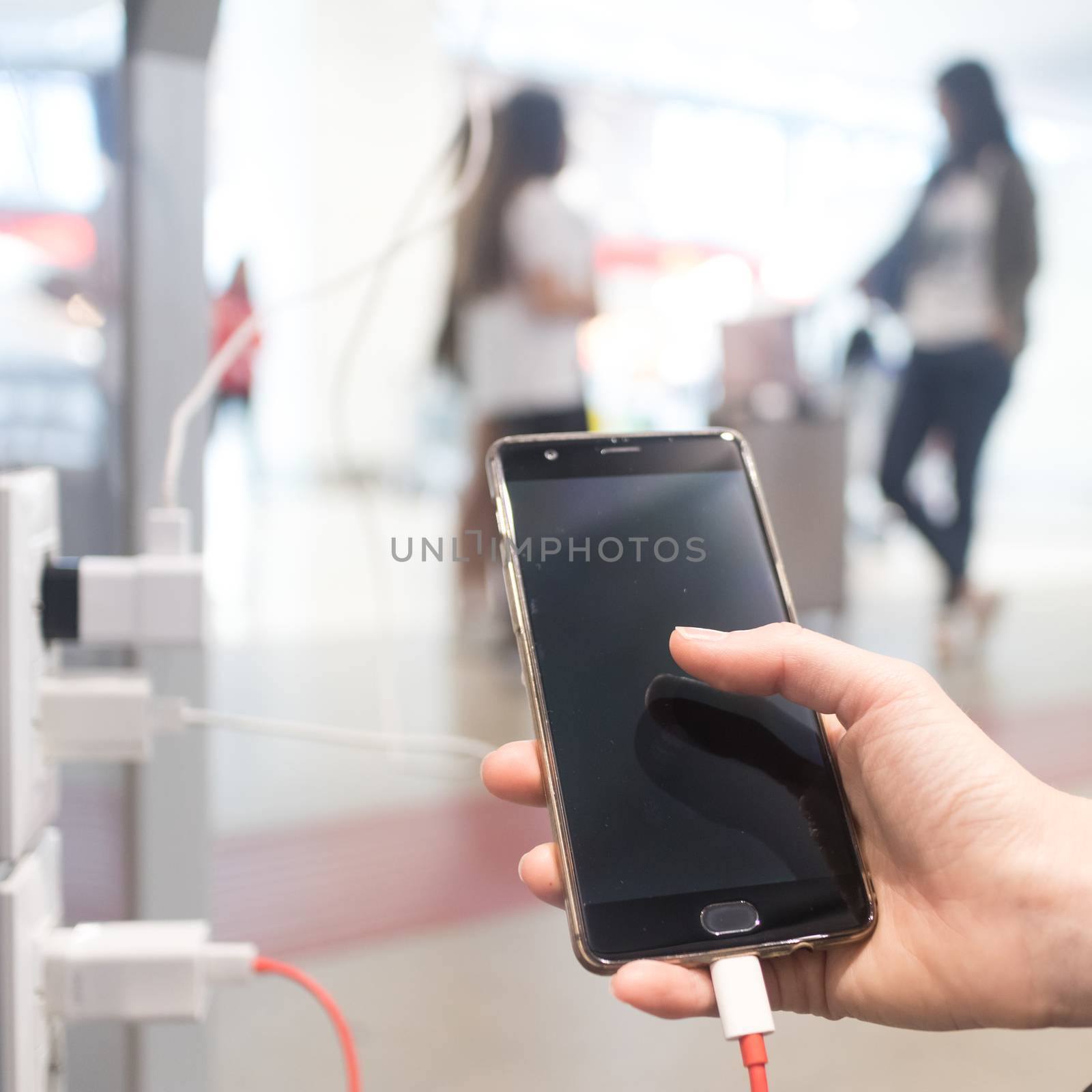 Female hands holding and using smartphone while charging it in a public place using electric plug and a charging cable.