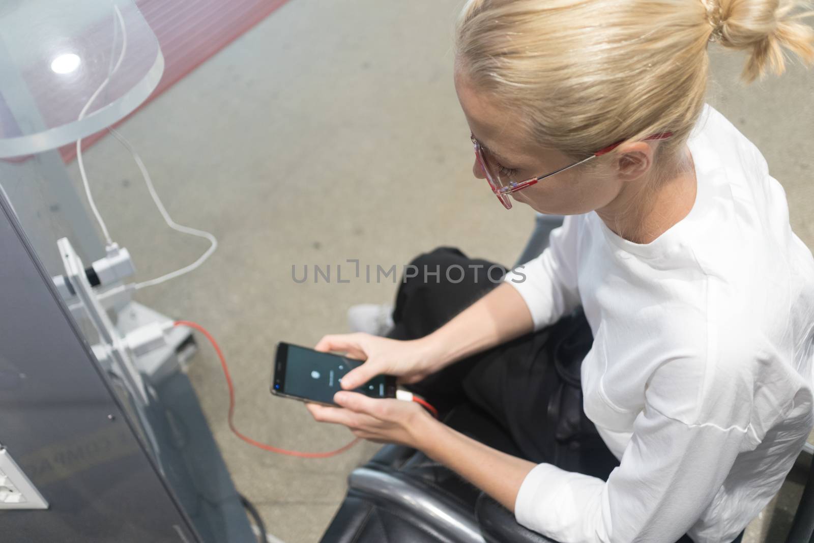 A young woman sitting at a charging station and looking at her smartphone. Recharging mobile phones from free charge station at the airport.