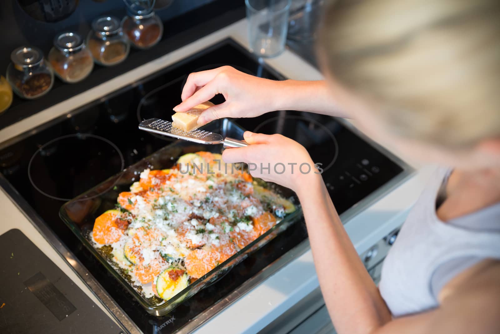 Female chef grinding parmesan cheese on row vegetarian dish ingredients in glass baking try before placing it into oven. Healthy home-cooked everyday vegetarian food.