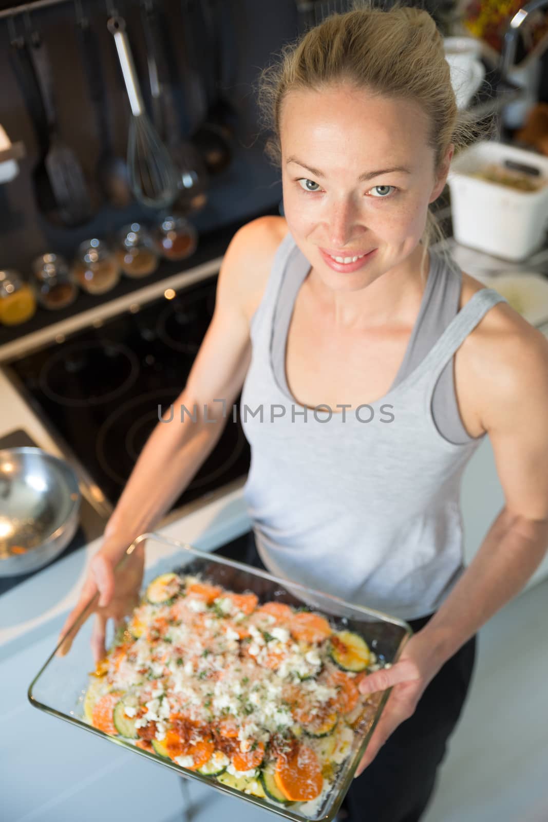Smiling young healthy woman holding and proudly showing glass baking try with row vegetarian dish ingredients before placing it into oven. Healthy home-cooked everyday vegetarian food.