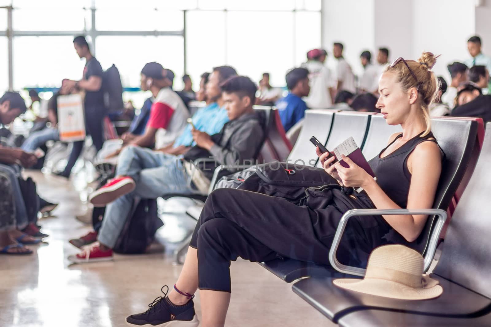 Casual sporty young blond female traveler using her cell phone while waiting to board a plane at the departure gates at the asian airport terminal.