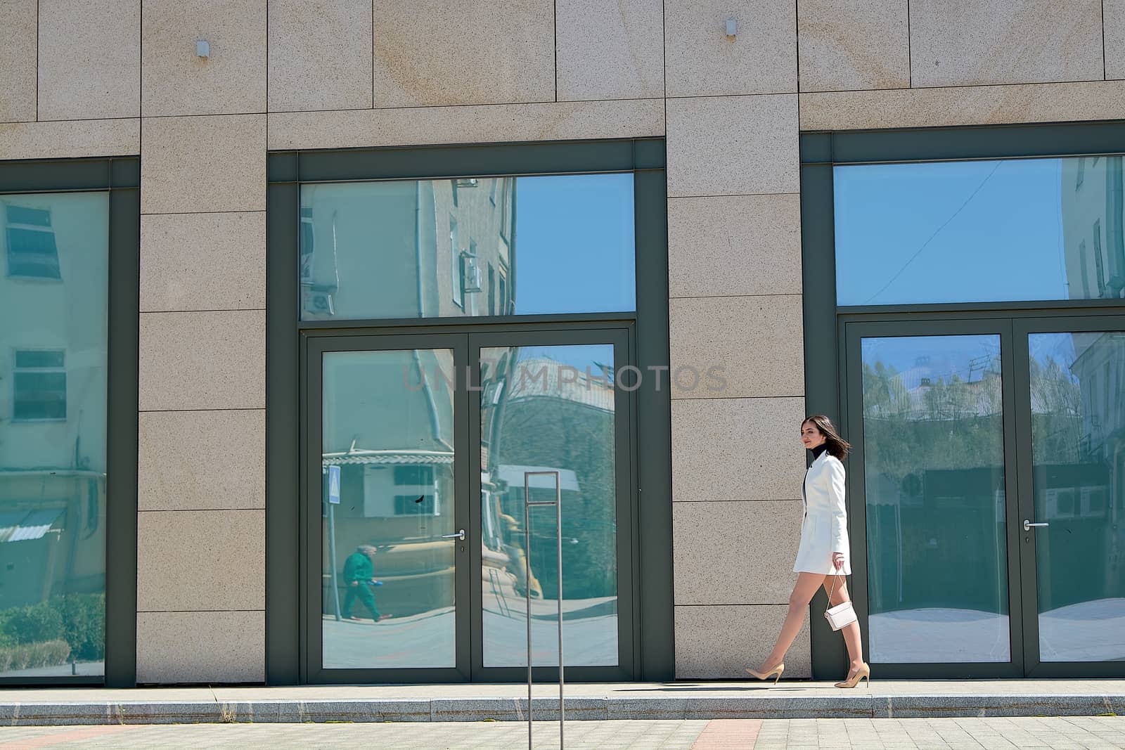 Elegant young woman dressed in a short white coat posing on a city street. Beautiful brunette woman. Modern urban woman portrait. Fashion business style clothes.