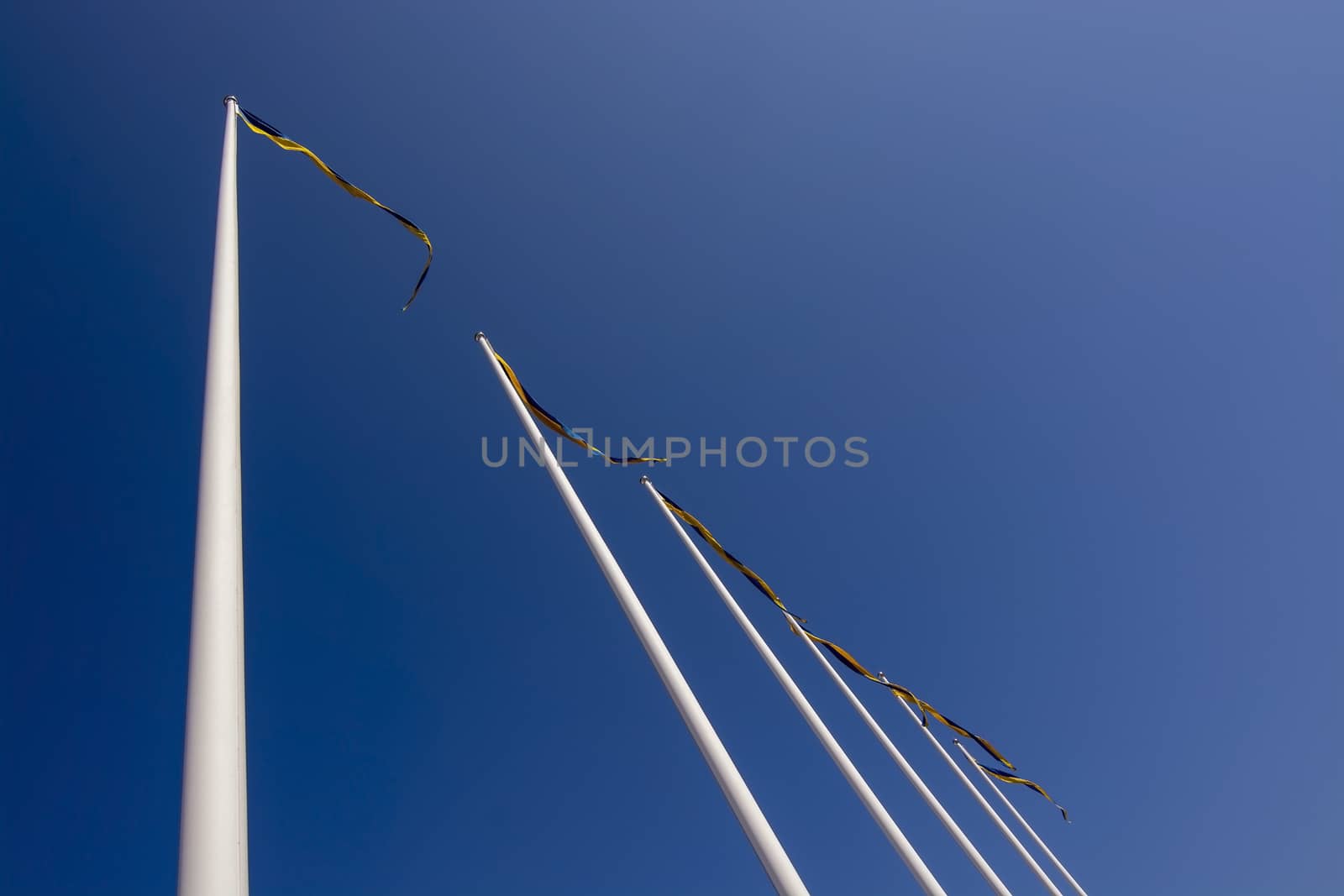 Swedish flagpoles in a diagonal row with blue and yellow pennants on a sunny day against blue sky.