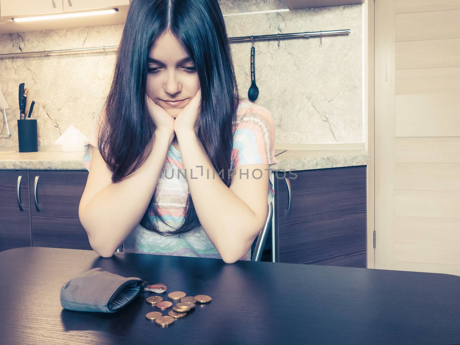 Concept of financial problems, a young disappointed woman with long dark hair, sits next to an old empty wallet with several coins on the table by galsand