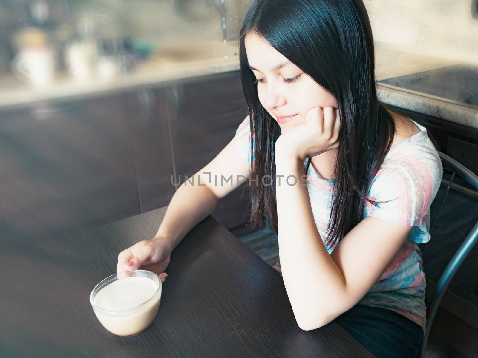 Young beautiful girl with dark long hair sits at a table with a cup of coffee and dreams about something.