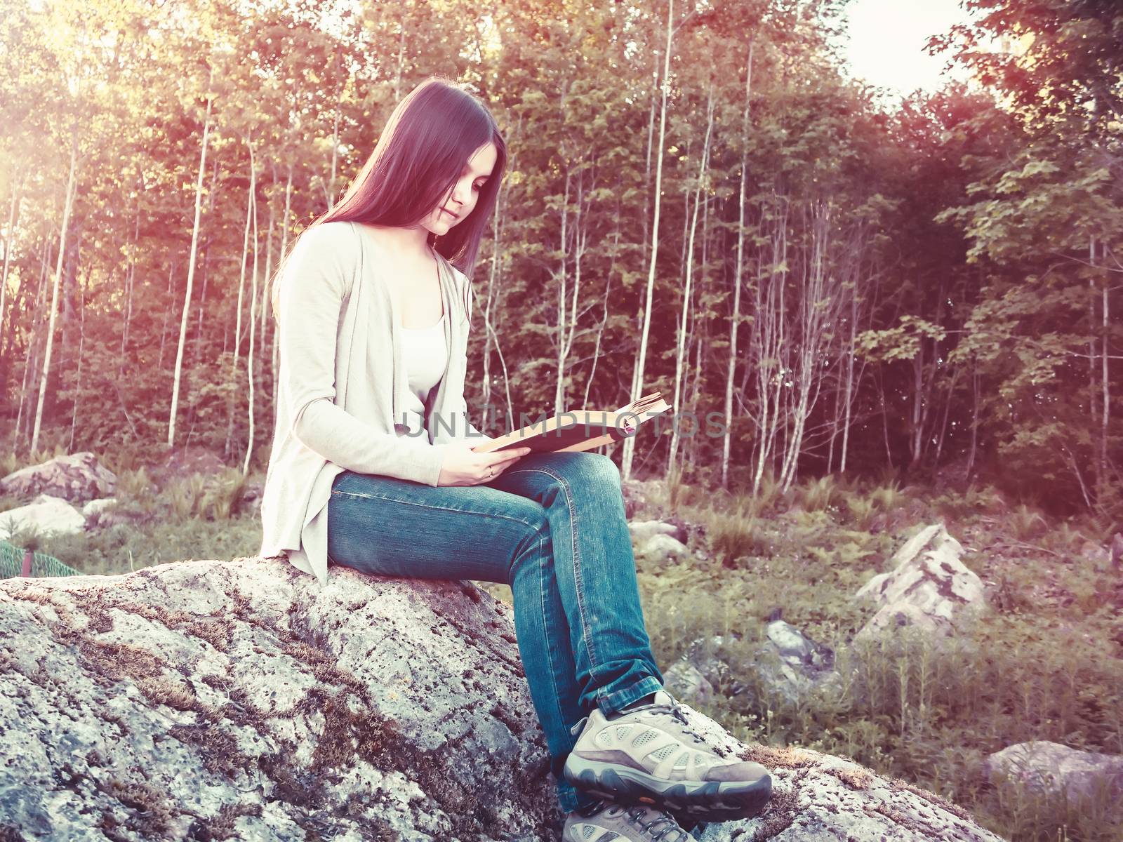 Young pretty girl reading a book sitting on a large rock in the forest by galsand