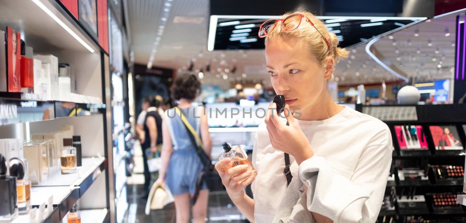 Beautiful blond young woman choosing perfume in retail store. Lady testing and buying cosmetics in beauty store.