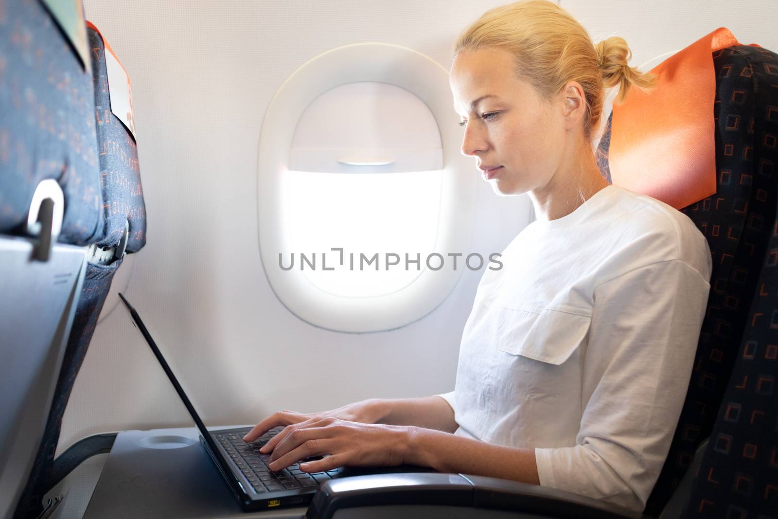 Attractive caucasian female passenger working at modern laptop computer using wireless connection on board of commercial airplane flight.