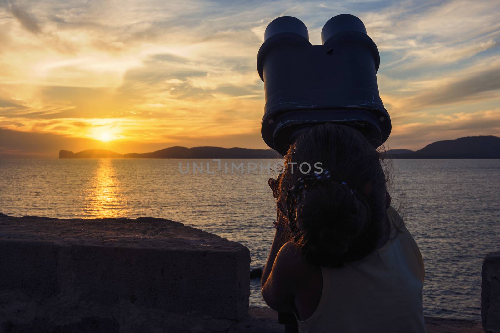 silhouette of a little girl looking through a binoculars for tourists during the sunset in front of the giant that sleeps in the Gulf of Alghero, Sardinia, Italy