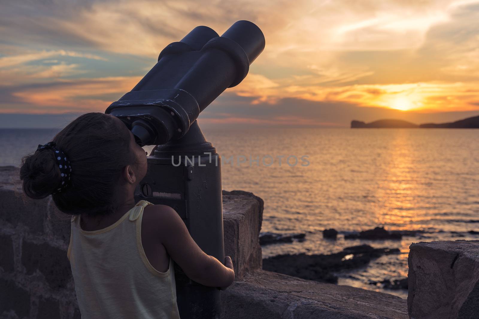 little girl looking through a binoculars for tourists during the sunset in front of the giant that sleeps in the Gulf of Alghero, Sardinia, Italy