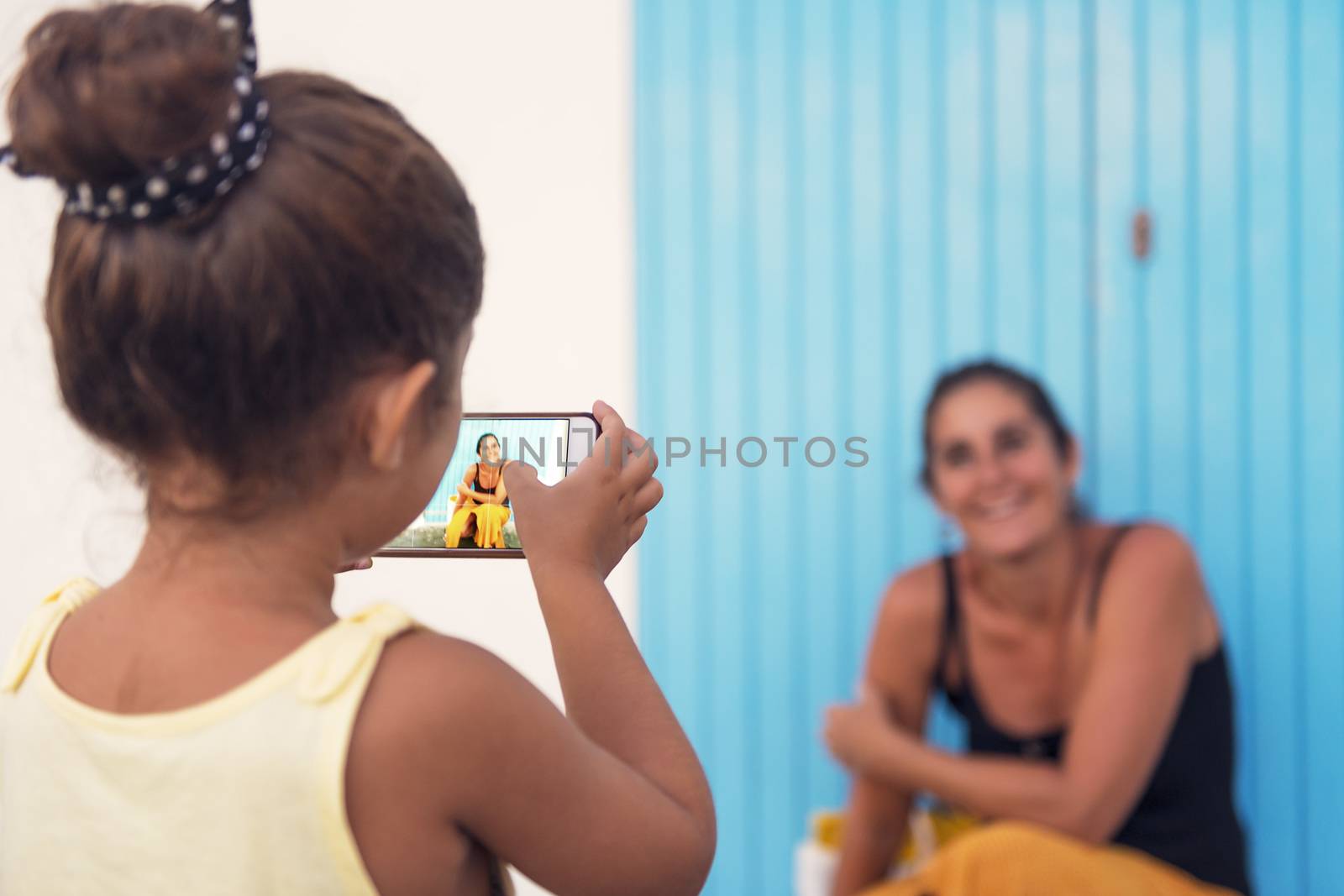 little girl taking photos with the smart phone to her mother in front of a blue door, single-parent family on vacation, divorced woman enjoying with her daughter