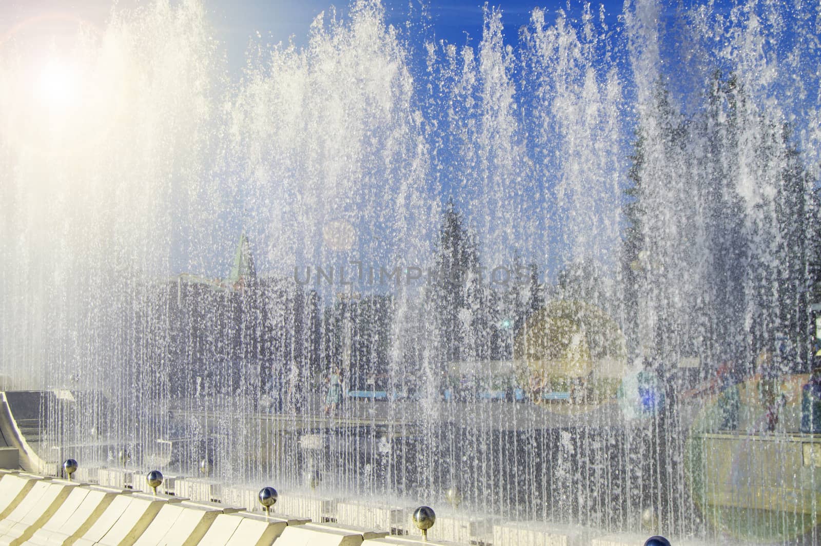 City fountain with water jets and random people, view through splashes on Sunny summer day.