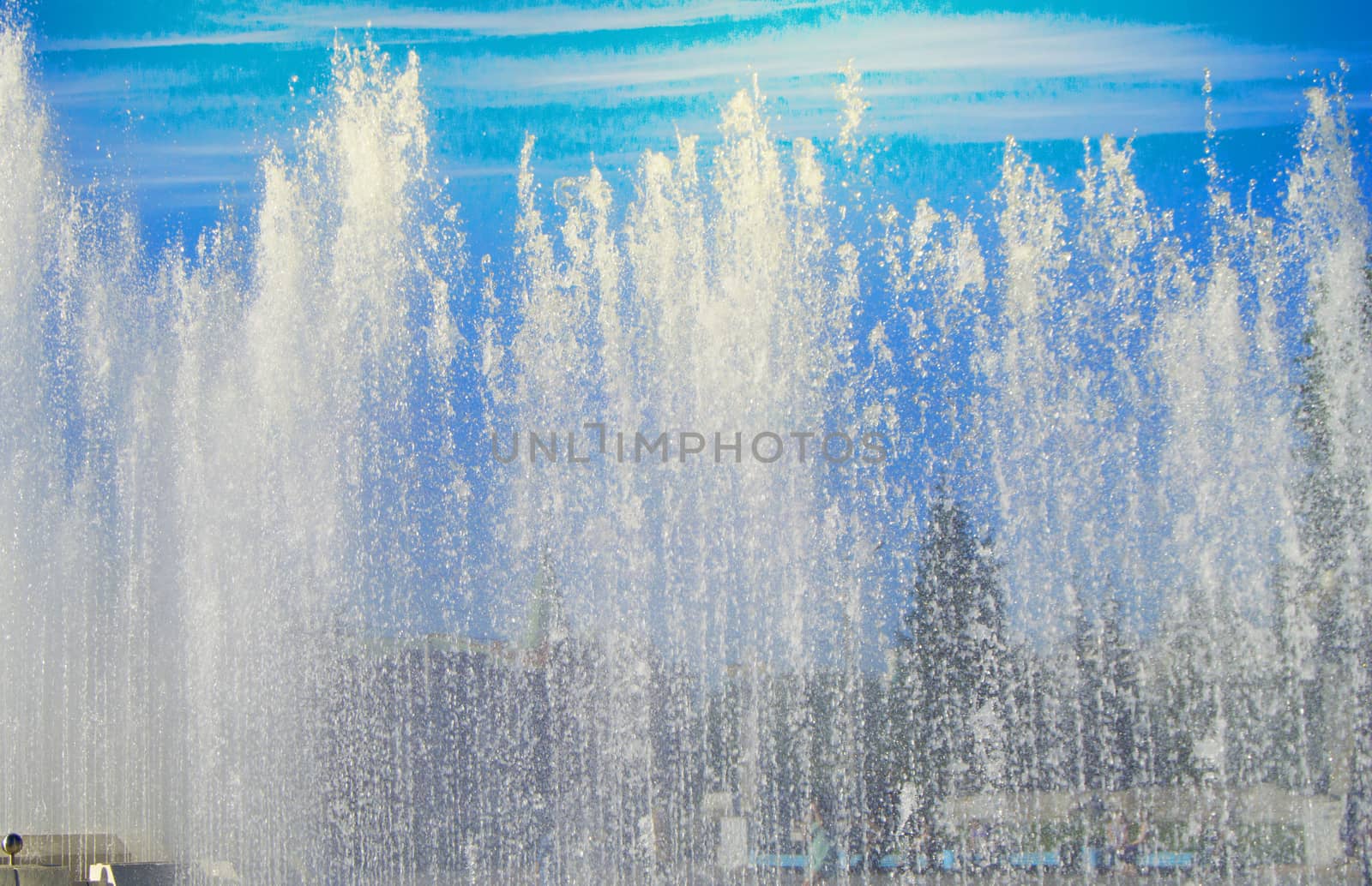 City fountain with water jets and random people, view through splashes on Sunny summer day.