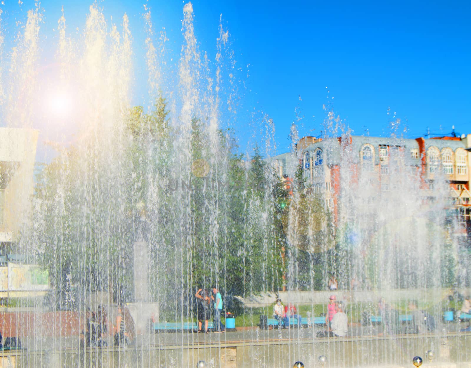 City fountain with water jets and random people, view through splashes on Sunny summer day.