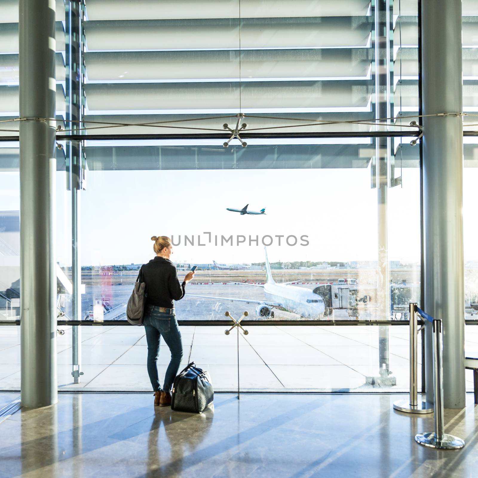 Young casual female traveler at airport, holding smart phone device, looking through the airport gate windows at planes on airport runway.