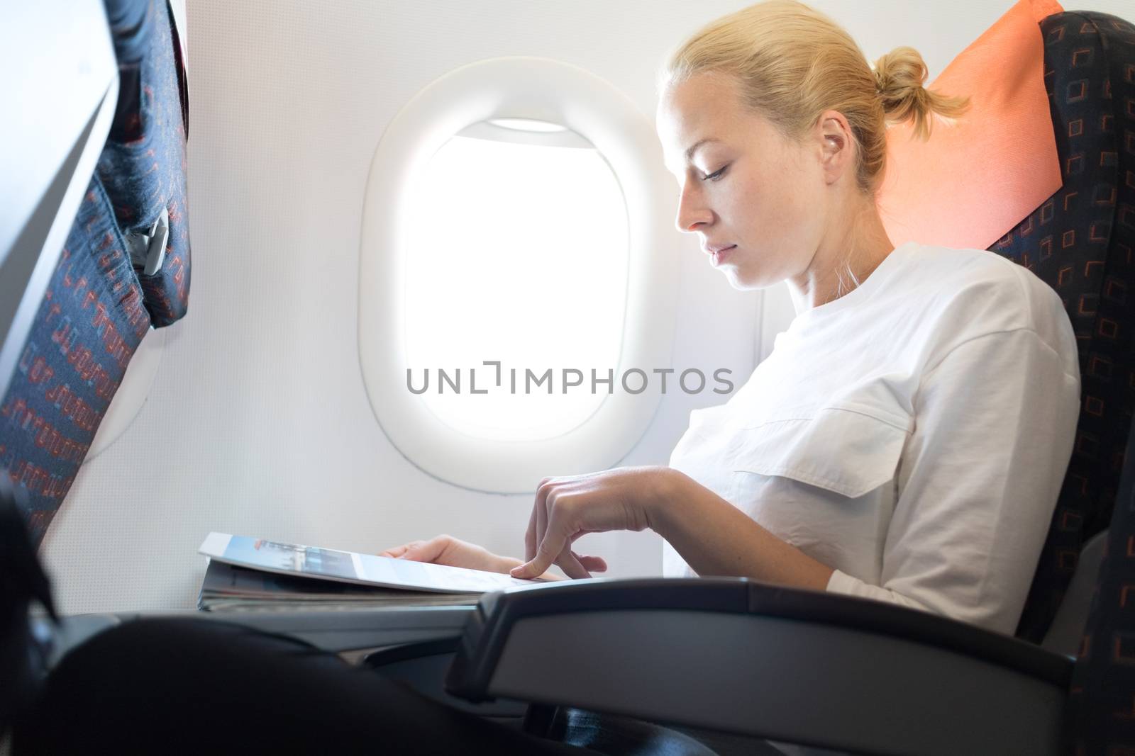 Woman reading in flight magazine on airplane. Female traveler reading seated in passanger cabin. Sun shining trough airplane window.