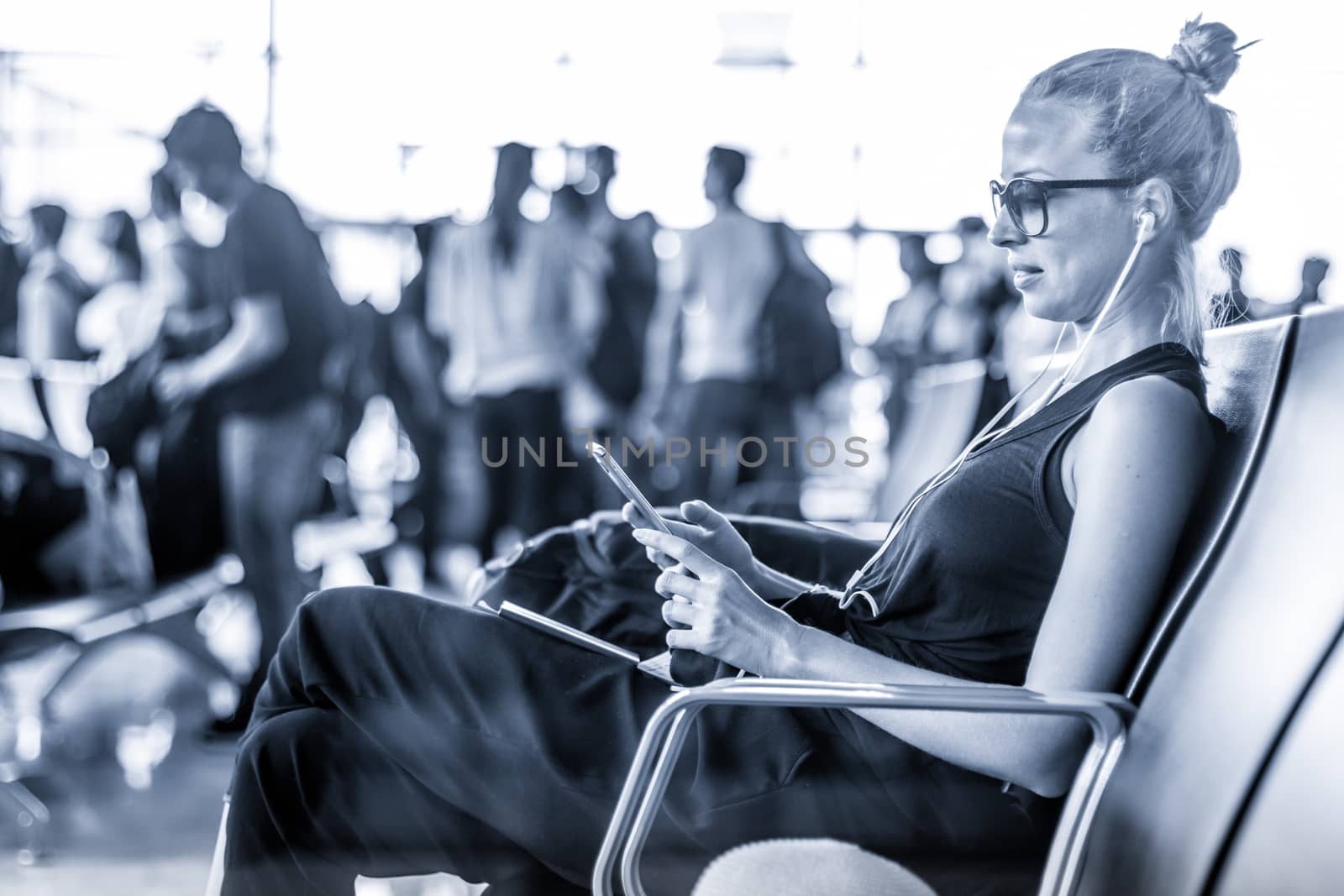 Casual sporty young blond female traveler using her cell phone while waiting to board a plane at the departure gates at the asian airport terminal. Blue toned black and white photo.