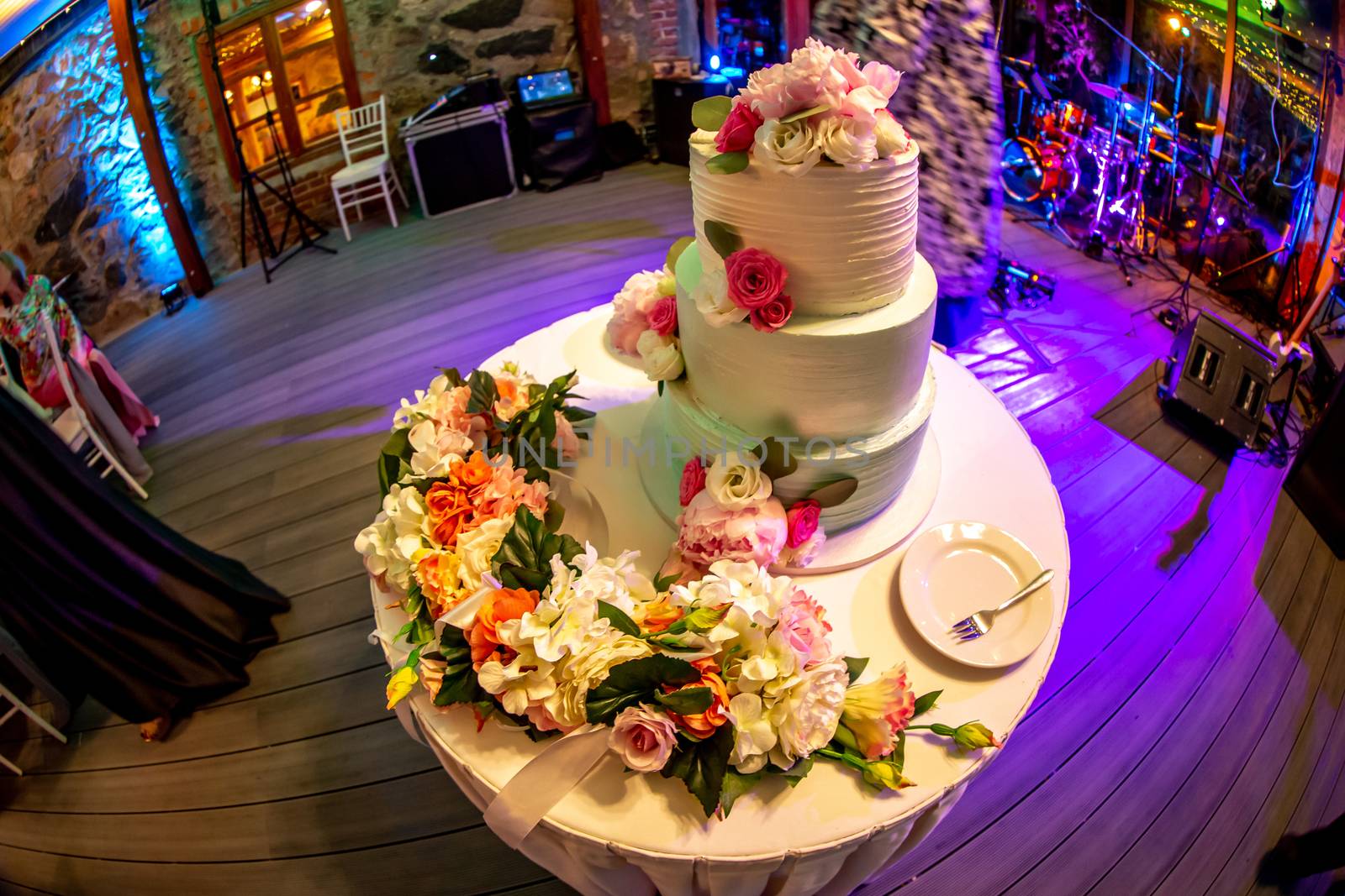 Wedding cake decorated with flowers on table. Three tiered wedding cake and plate with fork on white table. Shot with fisheye lens. 