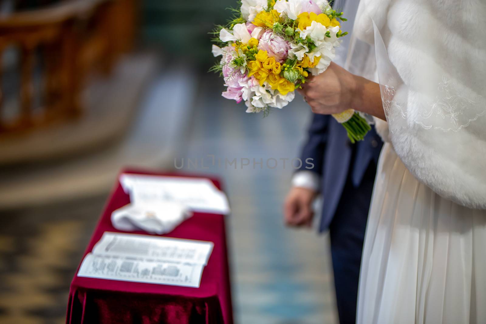 The bride holds a wedding bouquet of beautiful flowers in her hand. The bouquet consists of white, yellow and pink flowers. Marriage registration certificate and grom's hand is In the blur area. Bouquet of flowers in the hand of the bride during the marriage ceremony.