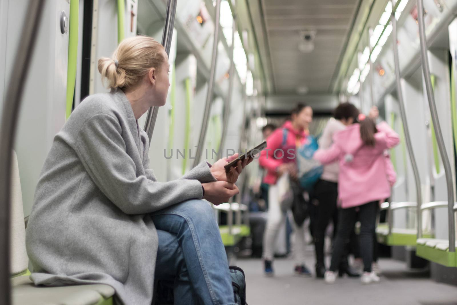 Beautiful blonde caucasian woman wearing winter coat and jeans using smart phone while traveling by metro. Public transportation concept.