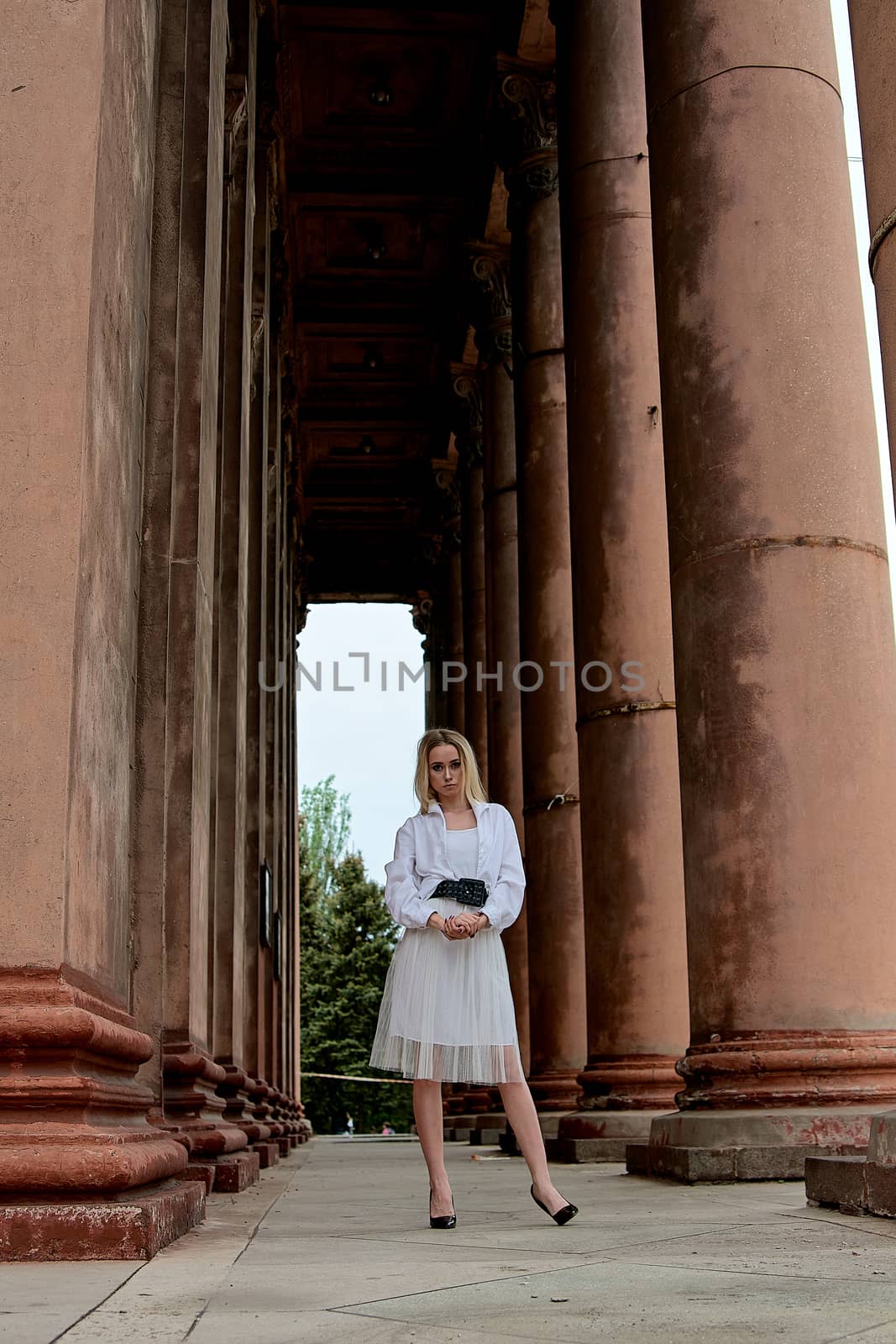 Fashion look's woman. Young woman modern portrait. Young woman dressed in white skirt and shirt posing near the old looking soviet union's architectural building with large pillars and bas-reliefs.