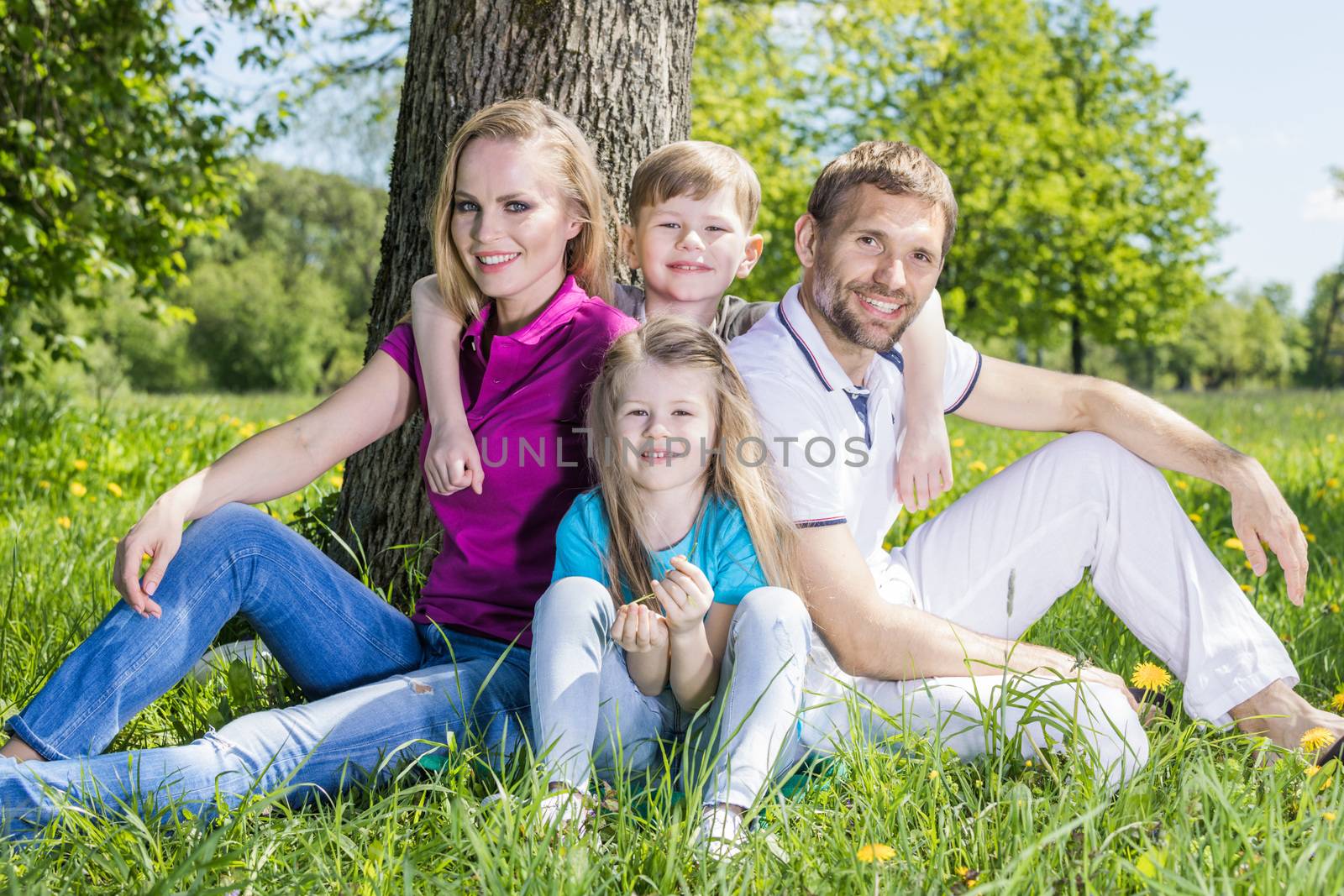 Parents and two children sitting under the tree on summer lawn. Happy family outdoors.