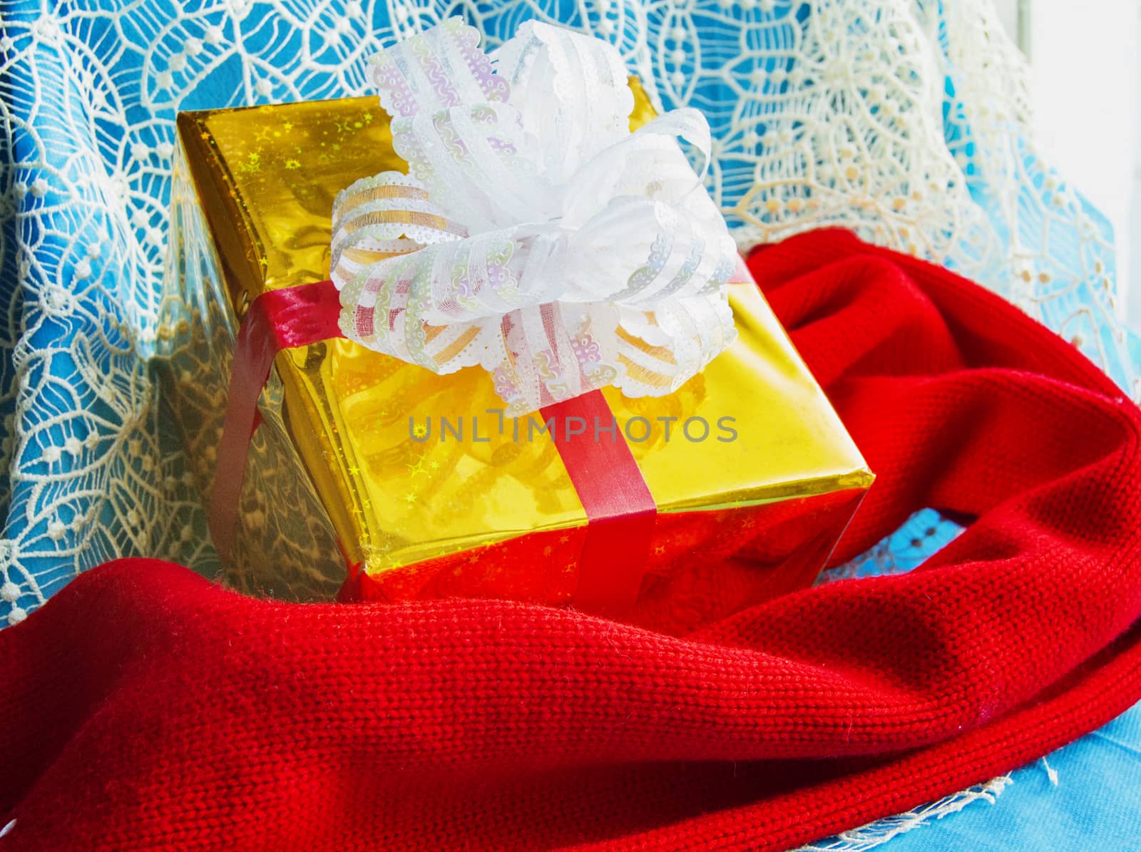 Gift box in gold foil package with white bow and red knitted scarf on beautiful blue Christmas background, Christmas layout concept by claire_lucia