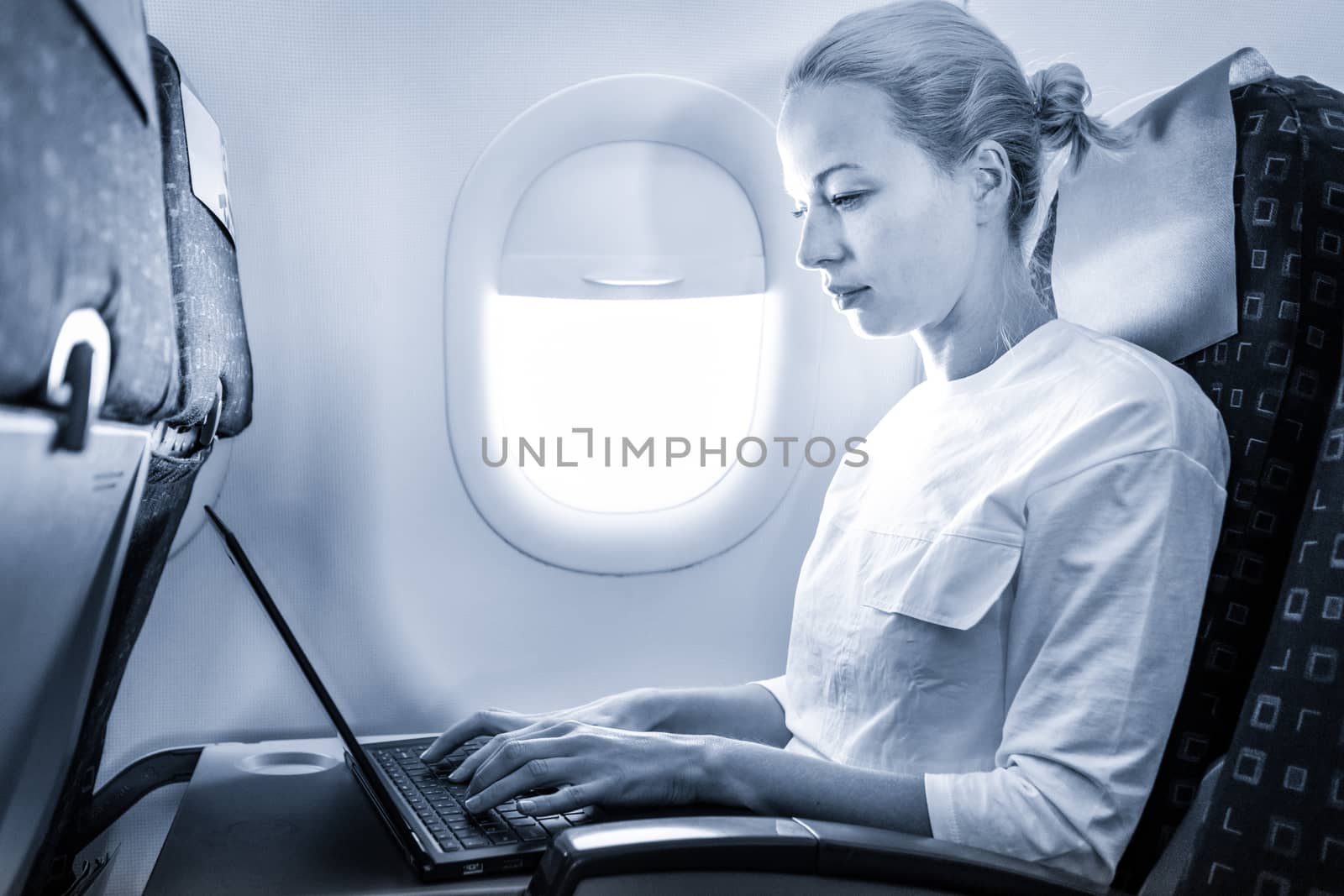 Attractive caucasian female passenger working at modern laptop computer using wireless connection on board of commercial airplane flight. Blue toned greyscale image.