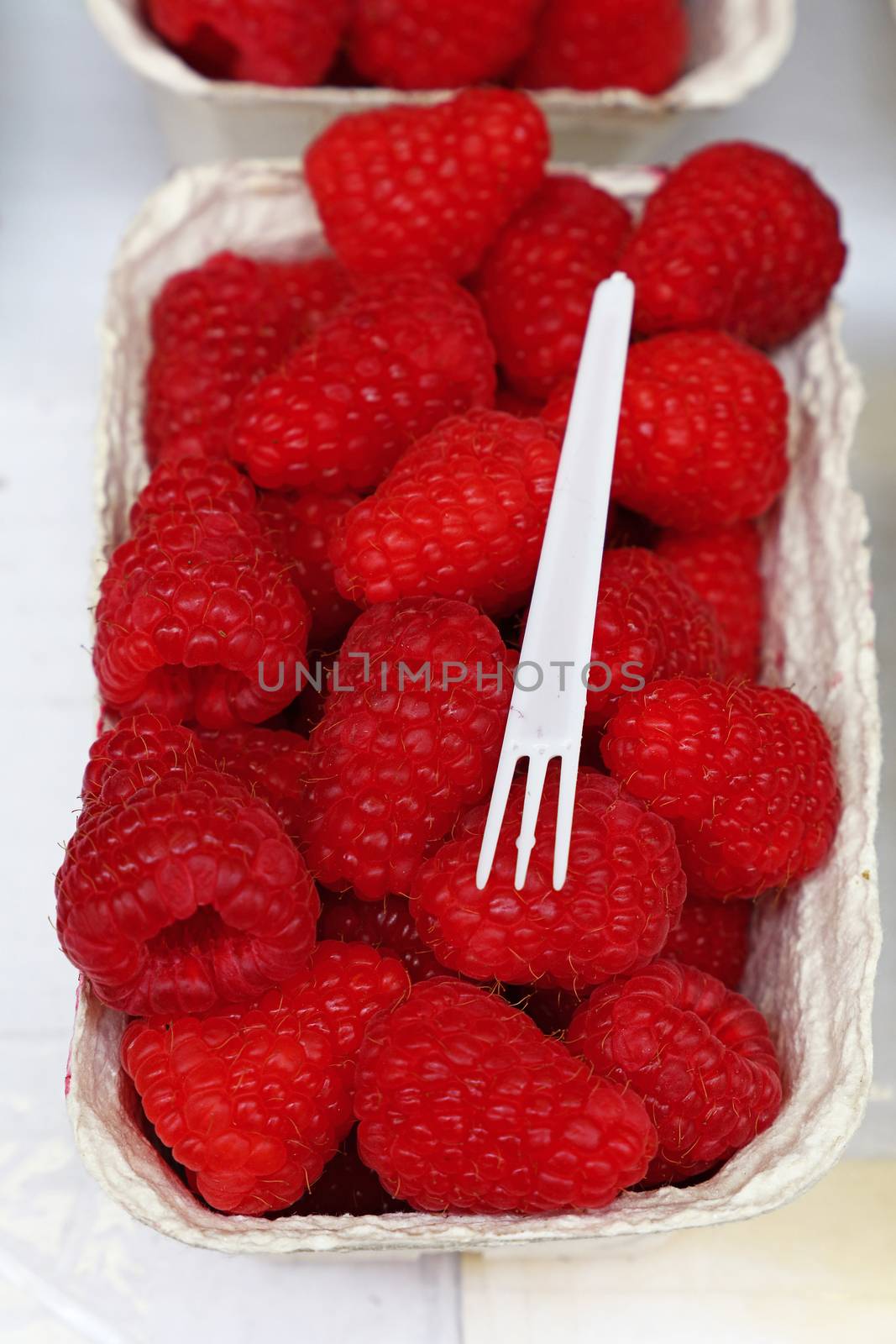 Close up fresh red raspberry berries in paper container on retail display of farmers market, high angle view