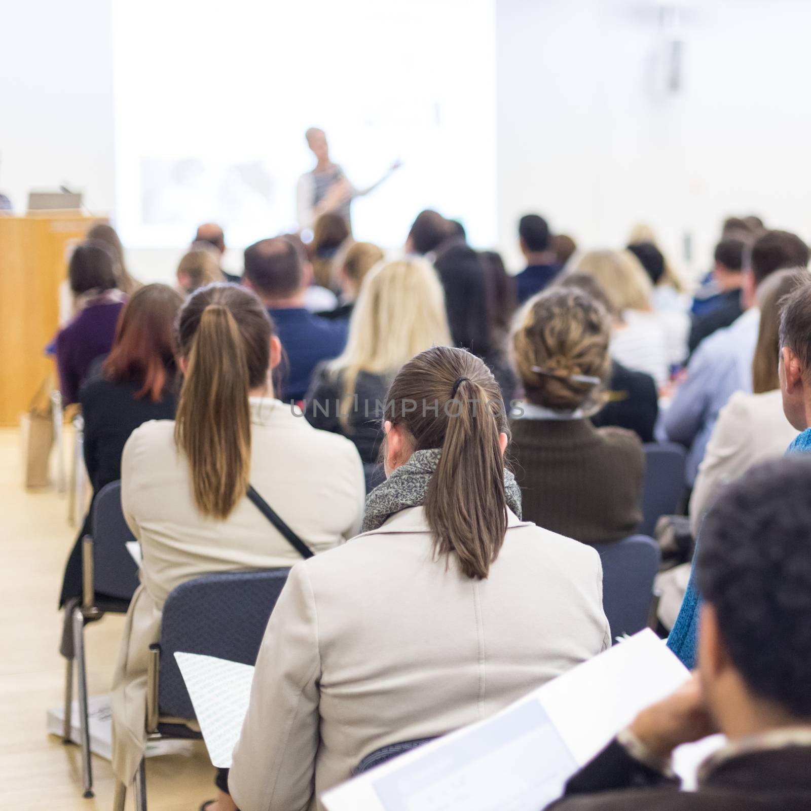 Business and entrepreneurship symposium. Female speaker giving a talk at business meeting. Audience in conference hall. Rear view of unrecognized participant in audience. Copy space on whitescreen.