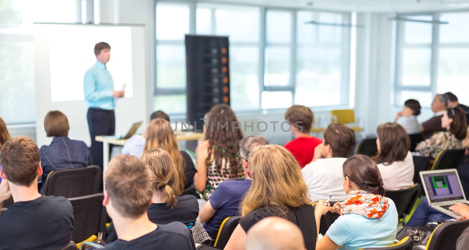 Business and entrepreneurship symposium. Speaker giving a talk at business meeting. Audience in conference hall. Rear view of unrecognized participant in audience.