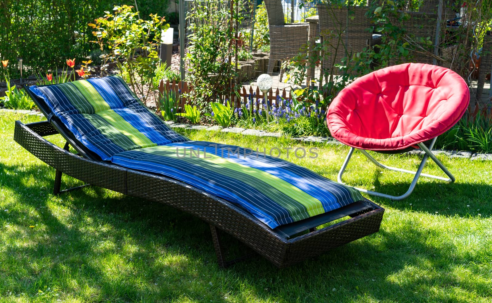 A blue and green sun lounger and a red folding moon chair in a garden in the sun. by daniel_albach