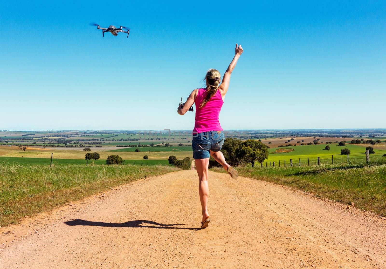 Woman leapiing for joy along a long dirt road  flying a drone.Views across a wide rural landscape of fields paddiocks and crops
