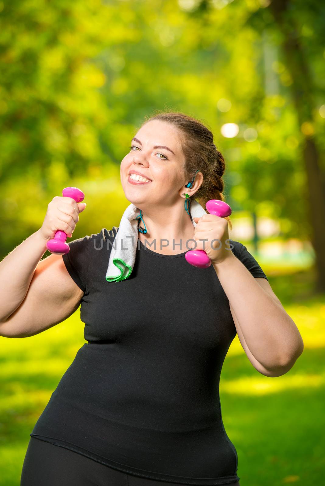 vertical portrait oversize woman in headphones with dumbbells in hand is engaged in sports in the park