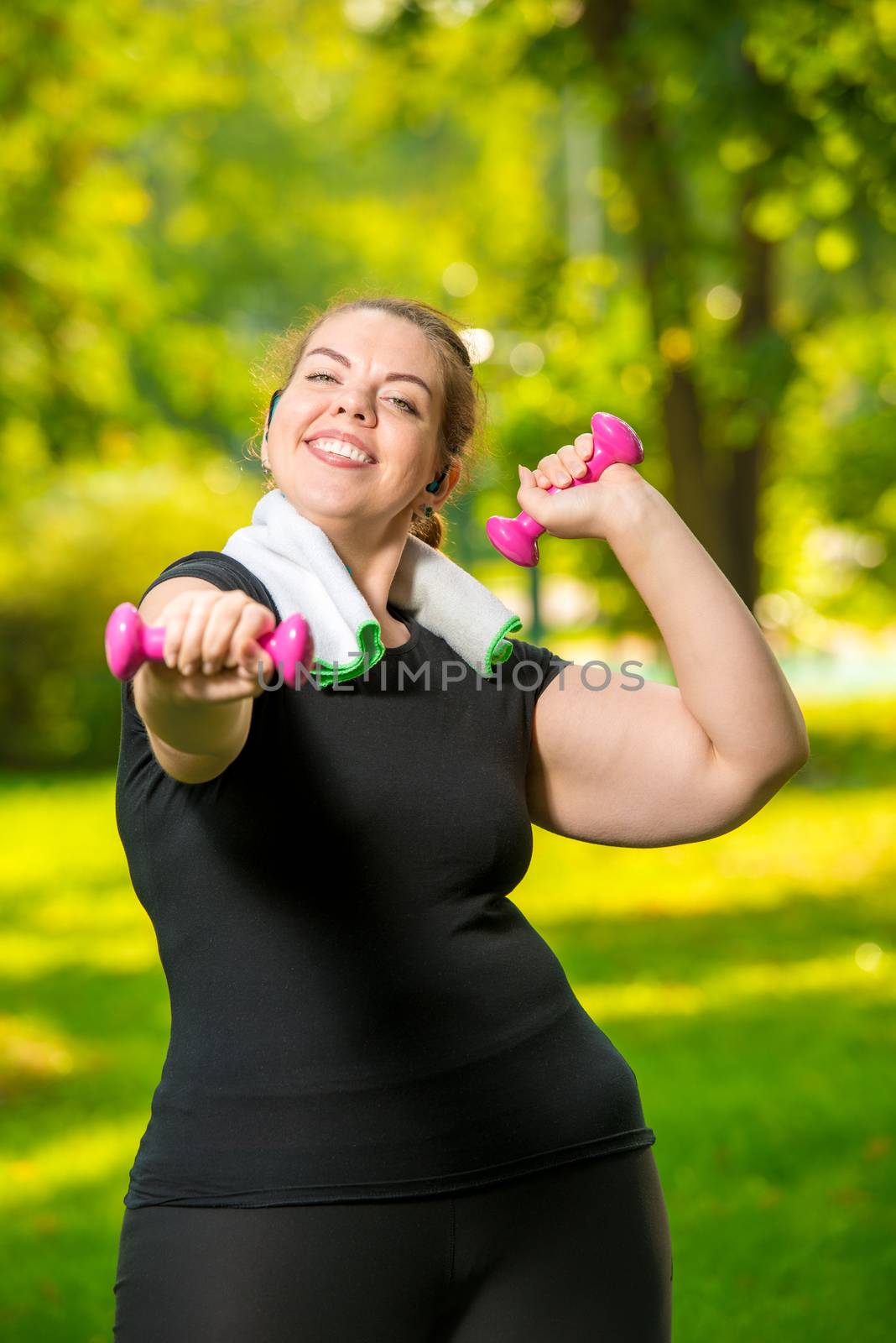 vertical portrait of a happy oversized woman in headphones with dumbbells in her hands playing sports in the park