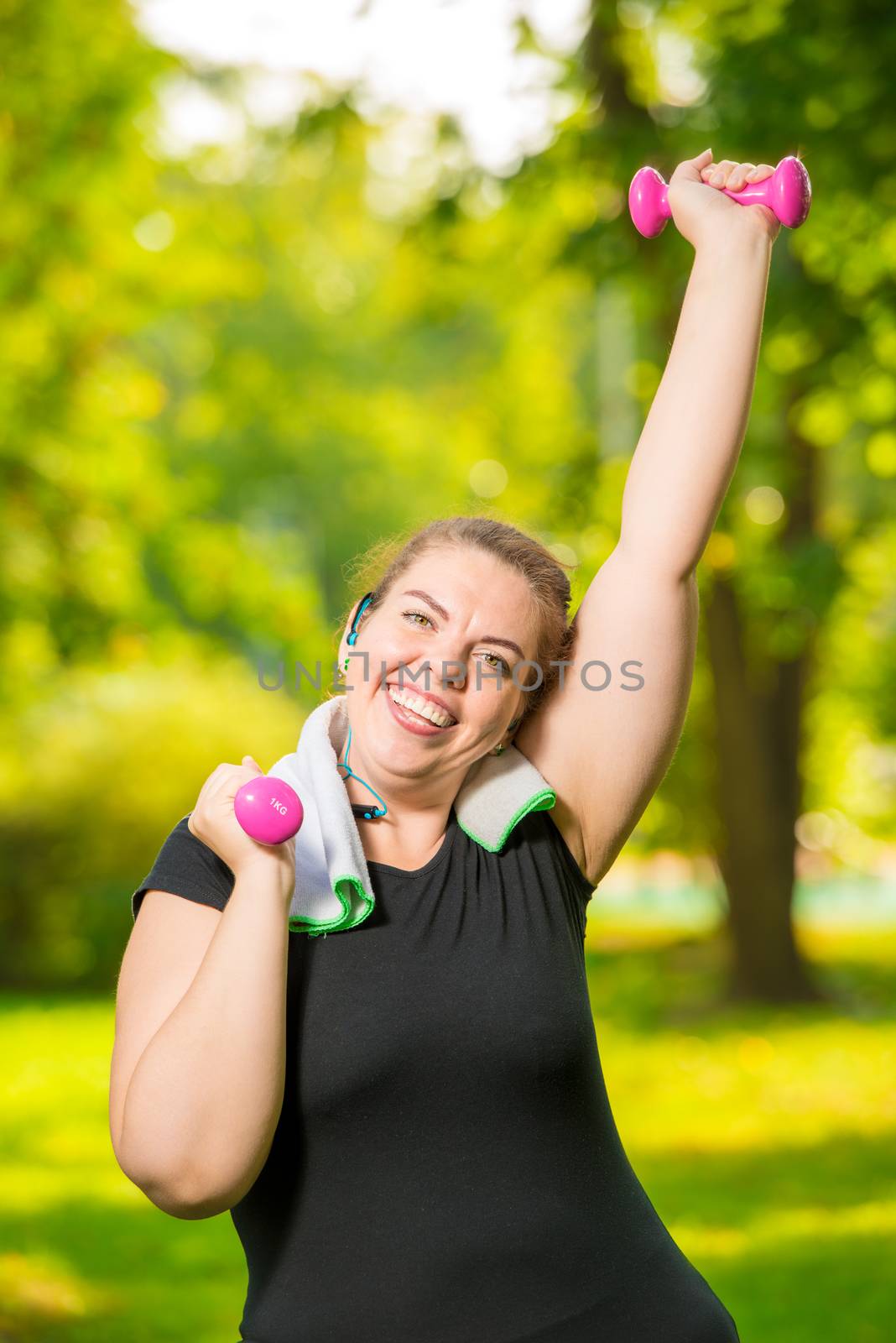 portrait of happy plus size model with dumbbells during workout in summer park