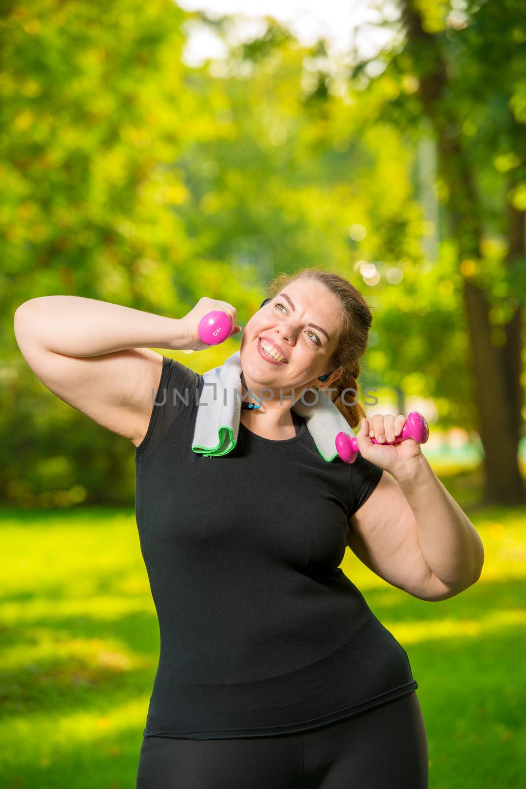 Vertical portrait of a happy plus-size model with dumbbells during a workout in a summer park