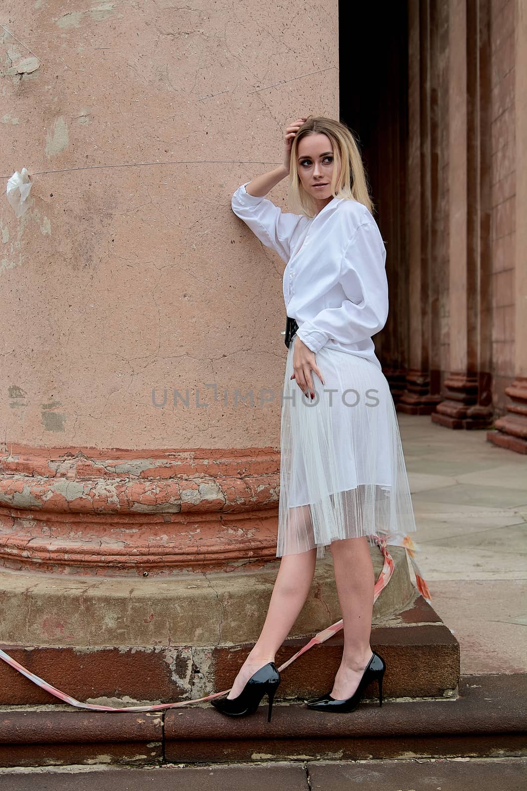 Young blonde woman dreses in white skirt and shirt posing on the steps of the old looking vintage building with large 
columns and bas-reliefs. Fashion look's woman. Young woman's modern portrait.