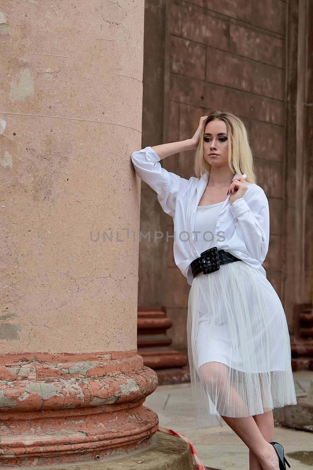Young blonde woman dreses in white skirt and shirt posing on the steps of the old looking vintage building with large 
columns and bas-reliefs. Fashion look's woman. Young woman's modern portrait.