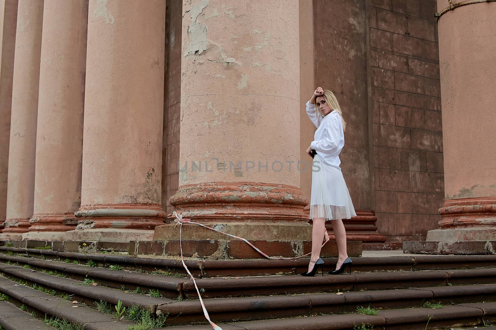 Young blonde woman dreses in white skirt and shirt posing on the steps of the old looking vintage building with large 
columns and bas-reliefs. Fashion look's woman. Young woman's modern portrait.